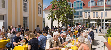 Menschen, die bei Sonnenschein an langen Tischen sitzen und gemeinsam essen
